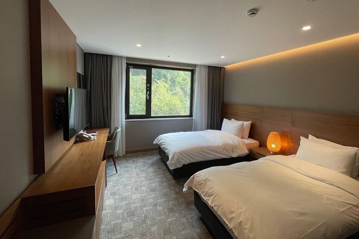 A hotel room with two beds, a desk with a flat-screen TV, a window with curtains, and a ceiling with recessed lighting.