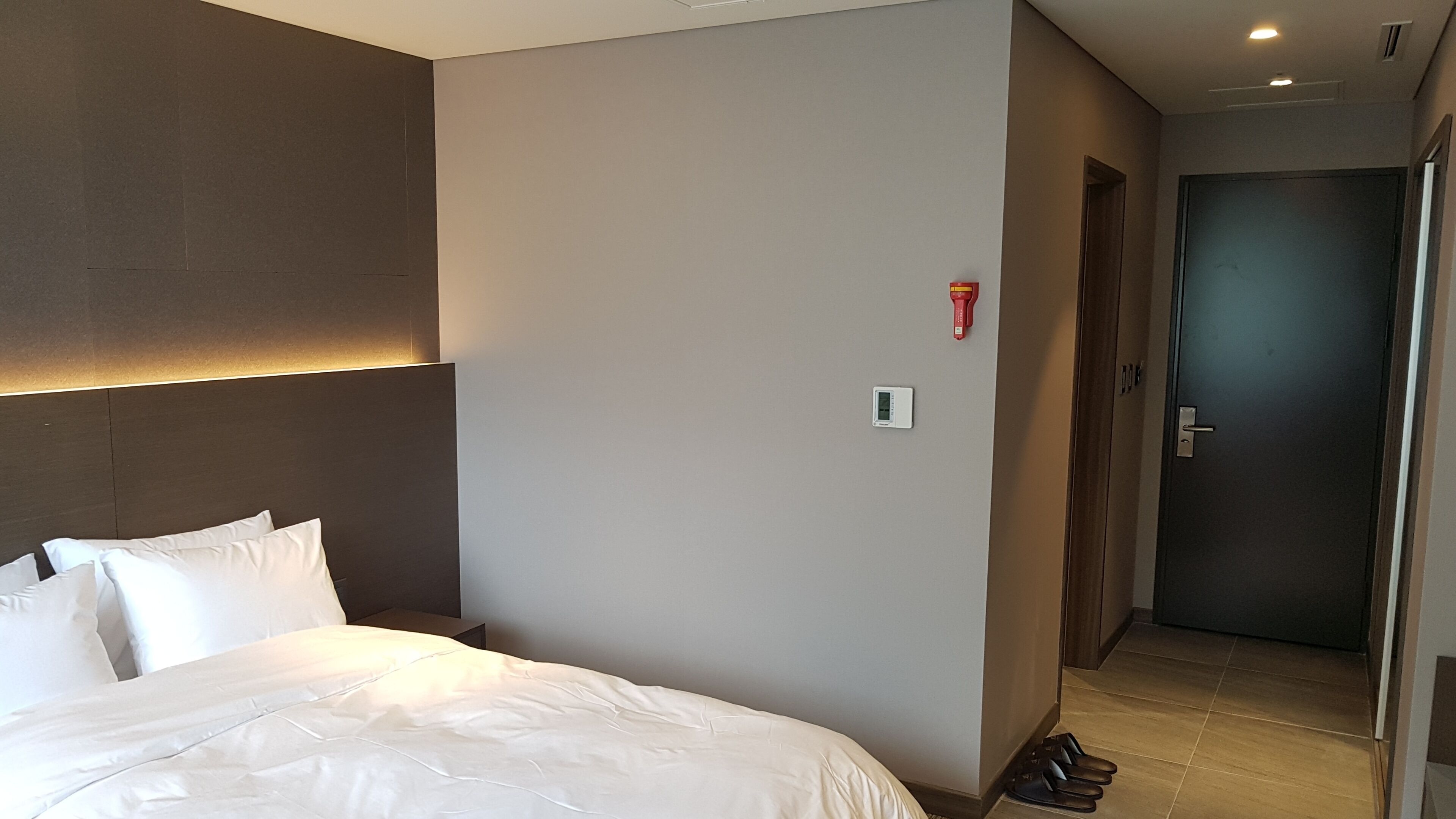 A hotel room with a bed, a nightstand, a wall-mounted thermostat, and a door leading to another room.