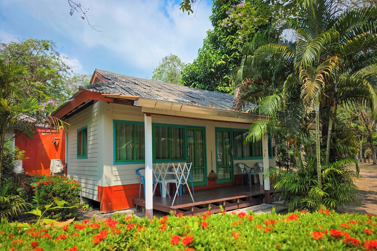 Ein kleines Haus mit Veranda, umgeben von üppiger Vegetation und blühenden Pflanzen.