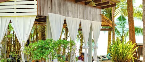 A rustic wooden structure with white curtains, outdoor seating, and potted plants.