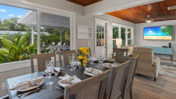 A dining area with a wooden table set for six, surrounded by chairs, with a view of greenery outside.