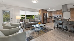 A modern kitchen with stainless steel appliances, a dining area with bar stools, and a living room with a sofa and armchairs.