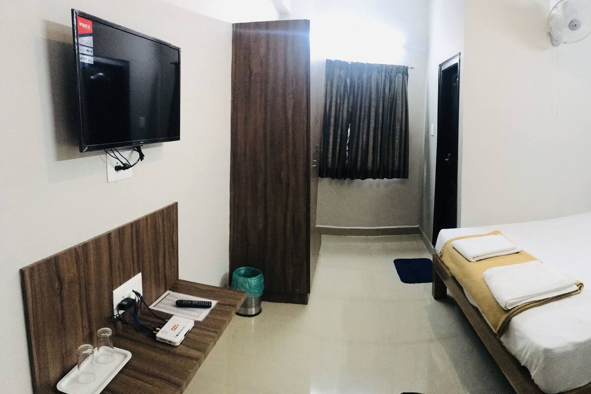 A hotel room with a bed, a desk with a chair, a TV mounted on the wall, and a window with curtains.