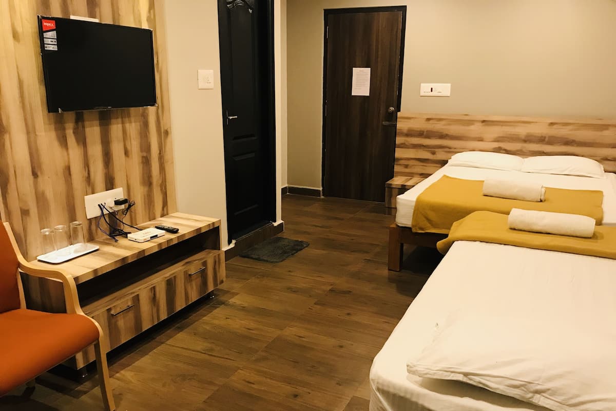 A hotel room with a wooden floor, a TV mounted on the wall, a desk with a computer and a remote control, and two beds with white bedding and yellow throw pillows.