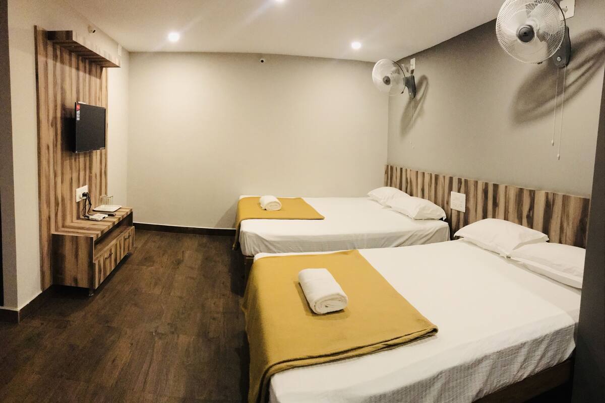 A hotel room with two beds, a wooden bench, a TV mounted on the wall, and a wall-mounted fan.