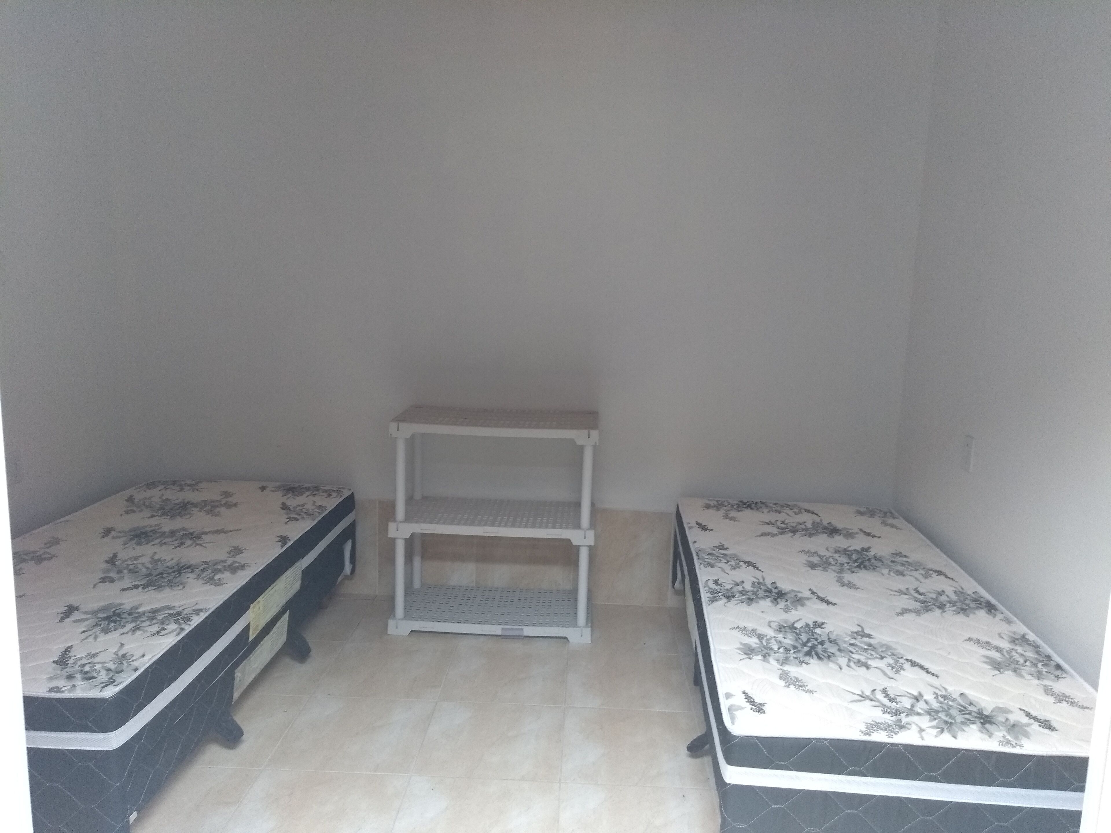 Um quarto pequeno e com pouca luz, com duas camas de solteiro, uma estante branca e piso de azulejos.