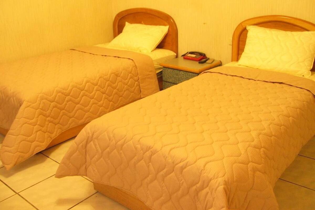 Duas camas de solteiro com cabeceiras de madeira e colchas combinando, uma mesa de cabeceira com um livro e um telefone, e um chão revestido com azulejos.