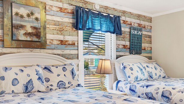 A bedroom with two beds, nautical-themed bedding, a coastal mural, and a window with a view of the outside.