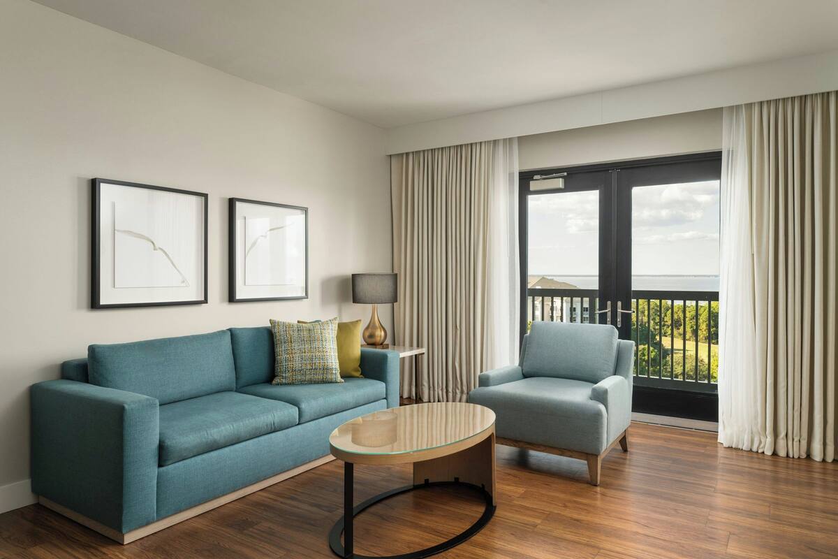 Un salon moderne avec un canapé de couleur turquoise, une table basse ronde et un fauteuil gris. Deux œuvres d’art encadrées ornent le mur, une lampe est posée sur une table d’appoint, et de grandes fenêtres offrent une vue sur l’extérieur.