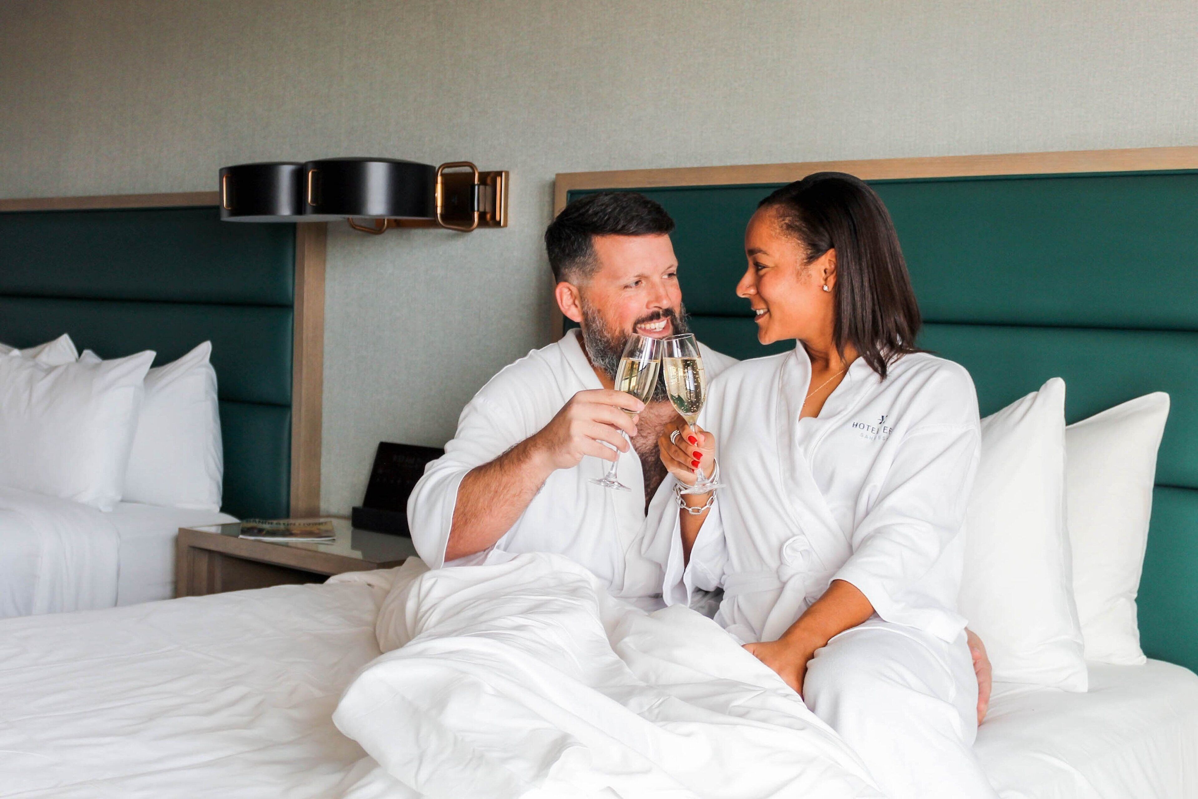 A couple in a hotel room with a green headboard, white bedding, and a bedside table with a lamp.