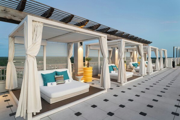 A rooftop lounge with white cushioned seating, white curtains, and a view of the sea.