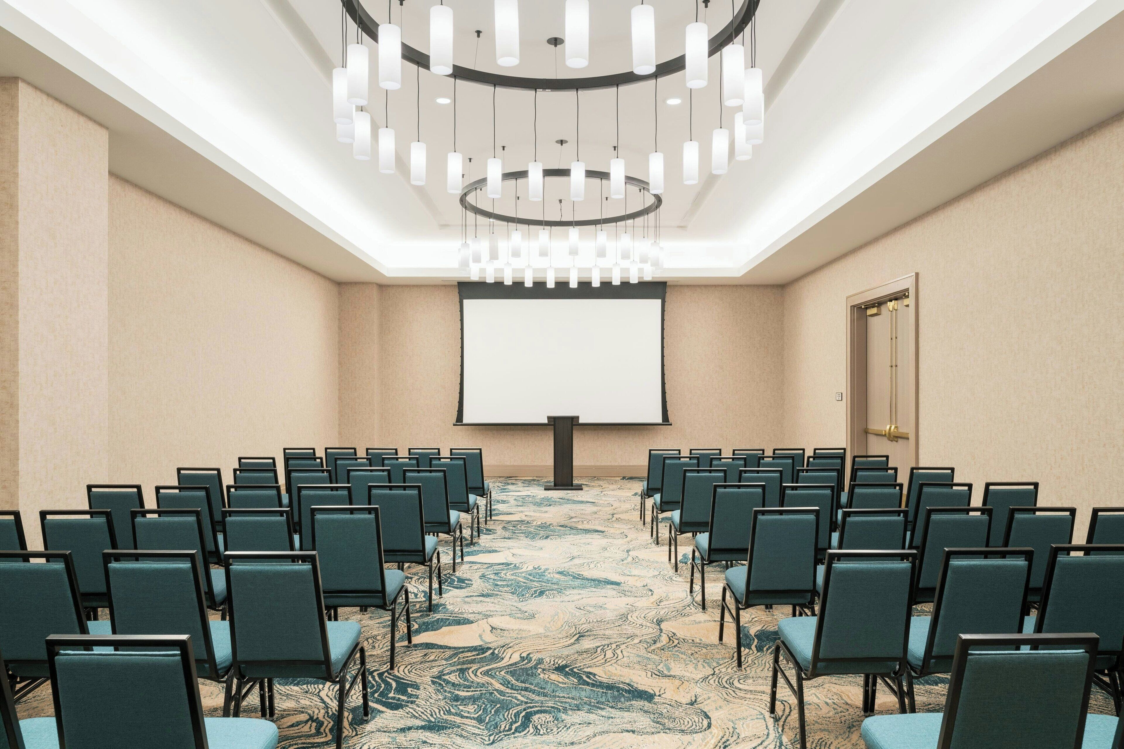 A conference room with rows of chairs facing a large screen, a patterned carpet, and a modern chandelier.