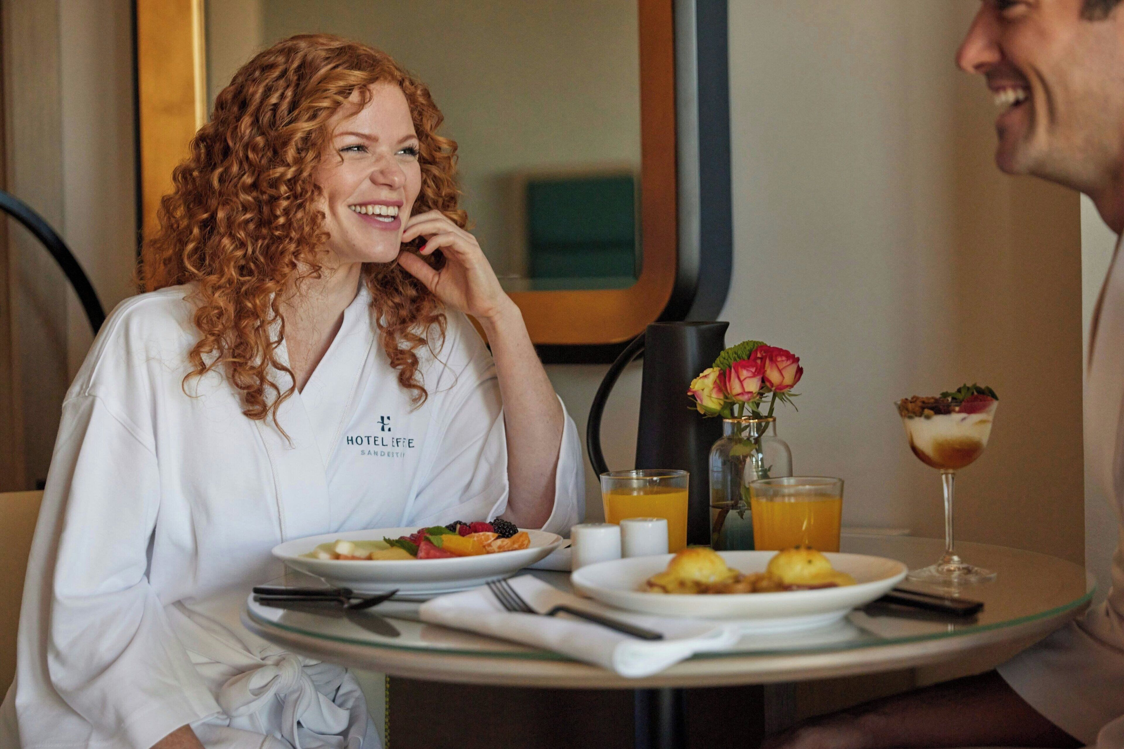 A hotel room with a dining table set for breakfast, including plates of food, a vase with flowers, and a glass of orange juice.