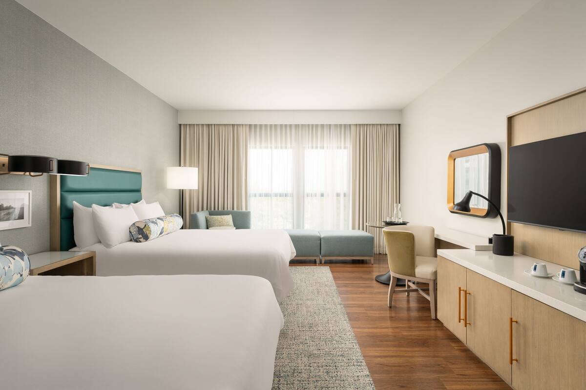 Une chambre d’hôtel avec deux lits, un bureau, une chaise, une télévision et une grande fenêtre avec des rideaux.