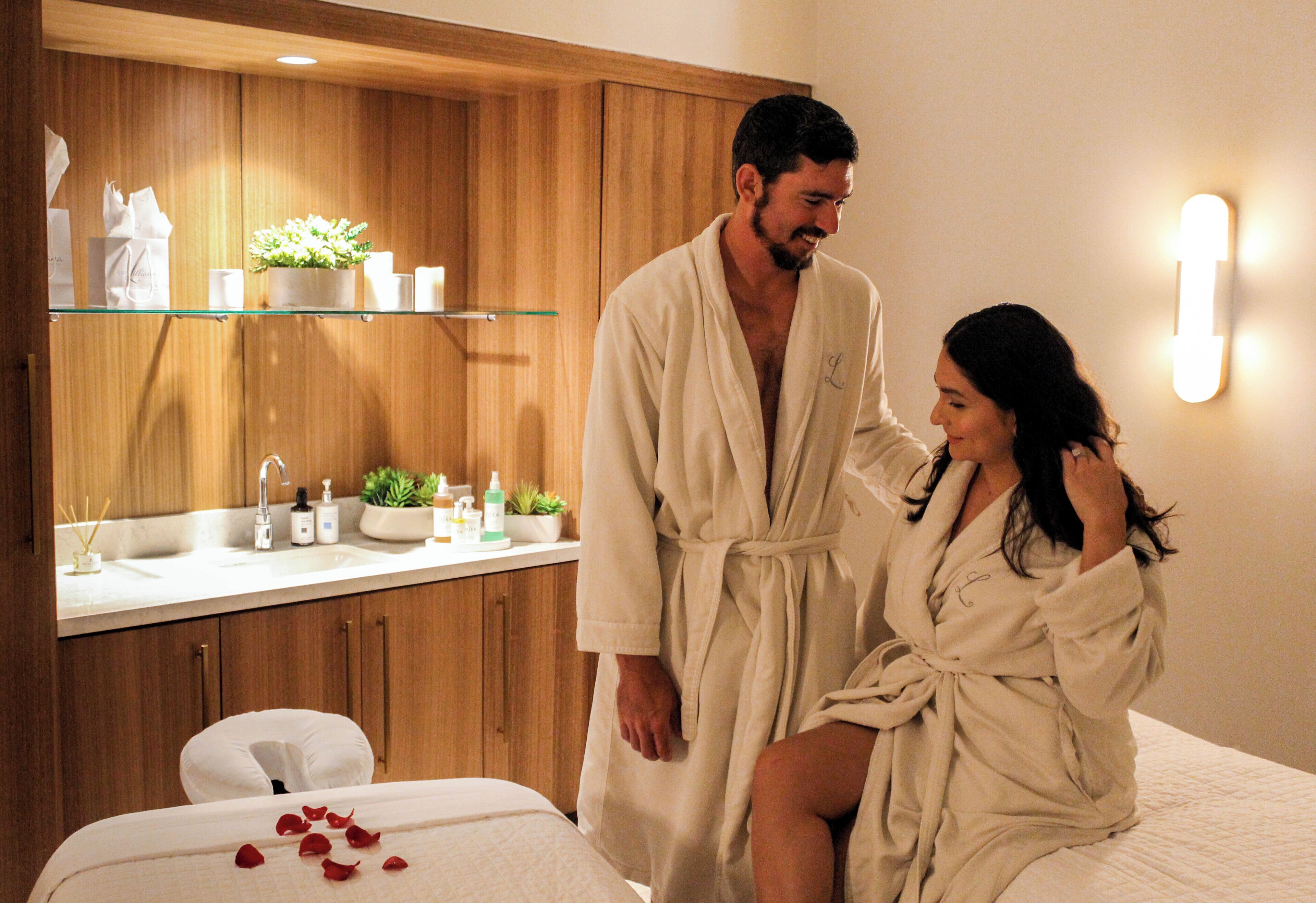 A couple in bathrobes in a hotel bathroom with a wooden vanity, a sink, and a mirror.