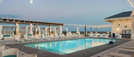 A rooftop pool area with lounge chairs and umbrellas, string lights, and a view of the moon.