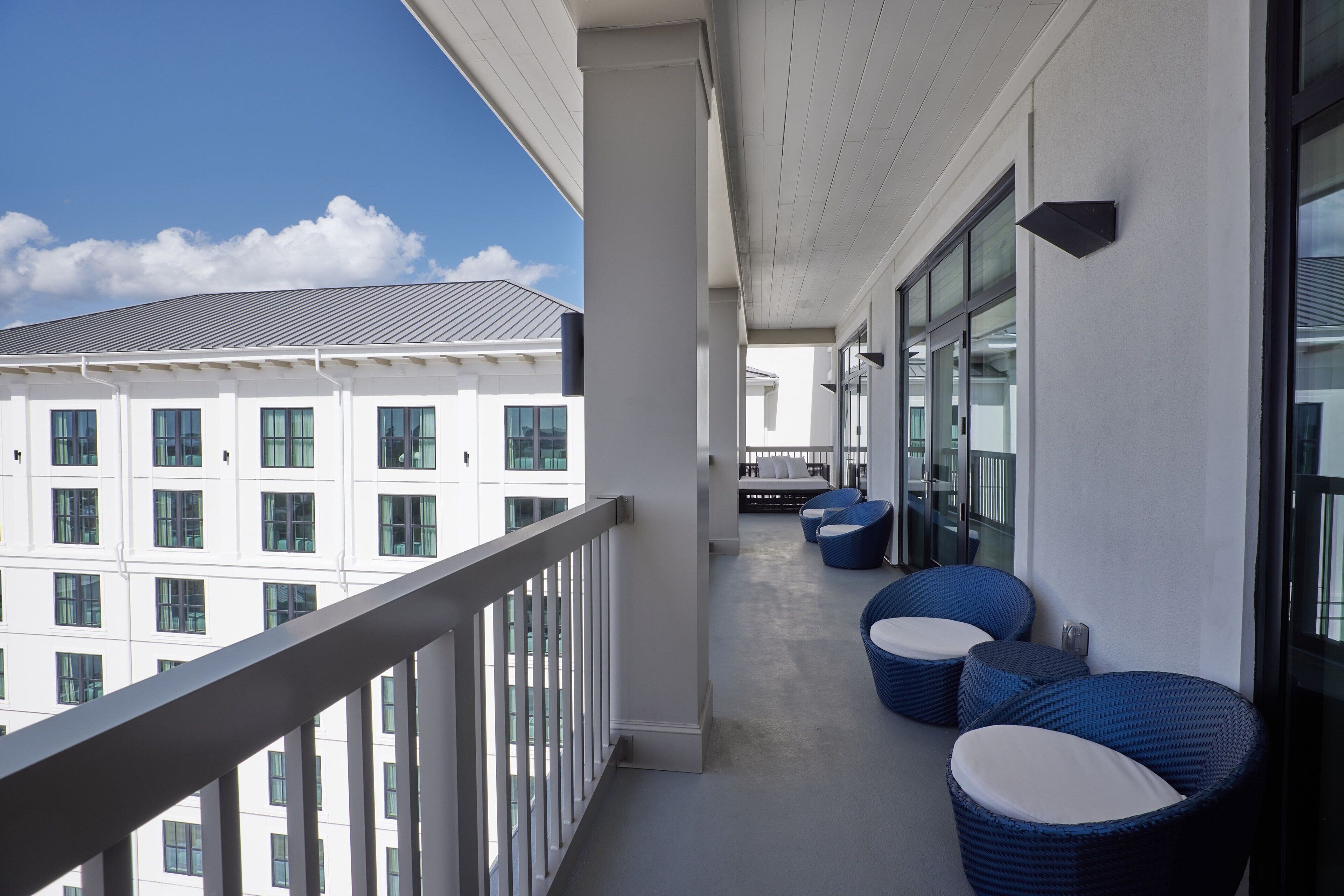 A balcony with a railing, a row of blue wicker chairs, and a view of a white building with multiple windows.