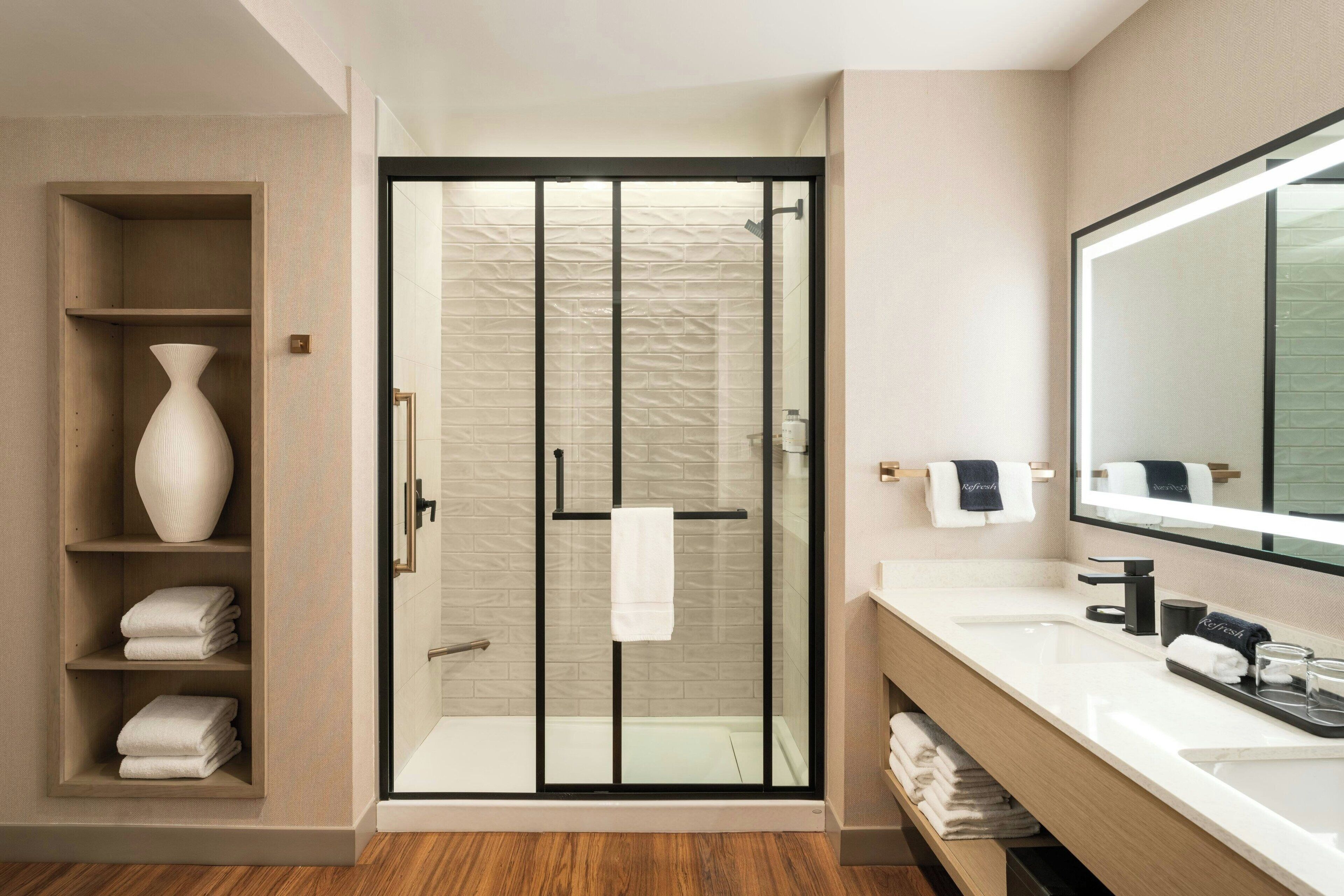 Une salle de bain moderne avec une douche à parois de verre, un lavabo blanc, un grand miroir et une étagère avec des serviettes pliées.