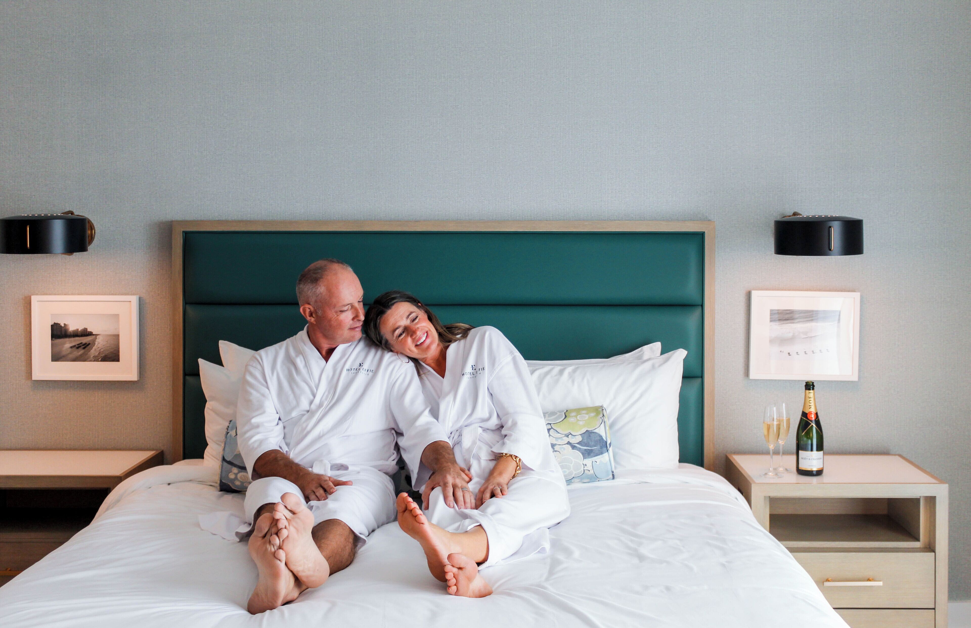 A couple in white robes on a bed with a teal headboard, two bedside tables with lamps, and a bottle of champagne on one table.