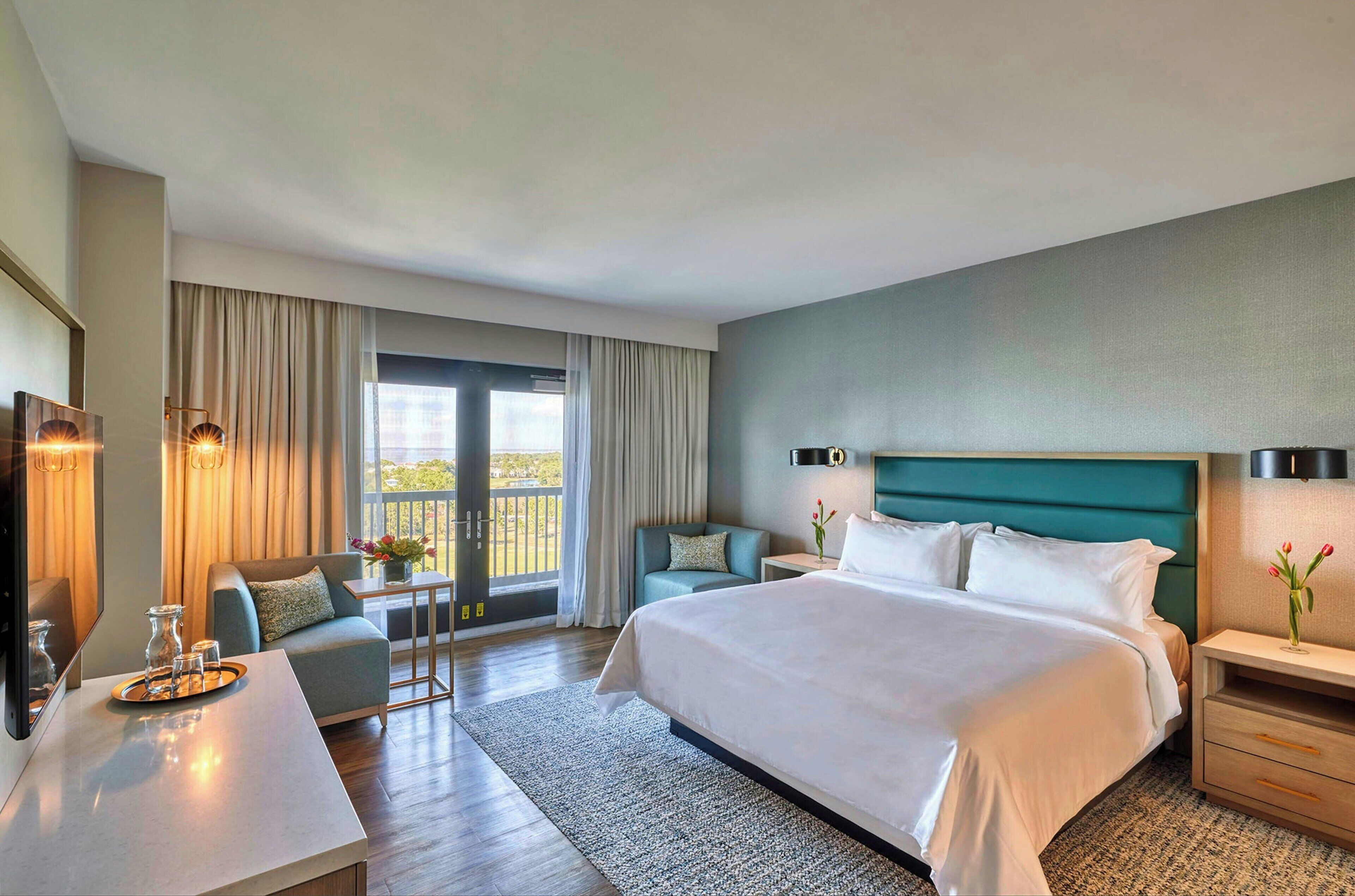 Una habitación de hotel moderna con una cama grande, una zona de estar con una mesa y sillas, un televisor y un balcón con cortinas.
