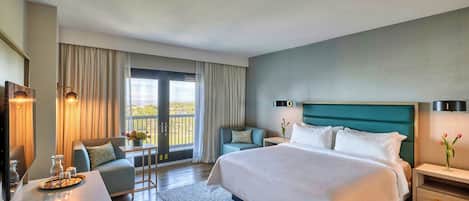 Una habitación de hotel moderna con una cama grande, una zona de estar con una mesa y sillas, un televisor y un balcón con cortinas.