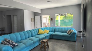 A modern living room with a blue sectional sofa, a wooden stool, and a large window offering a view of a green outdoor area. - Beautiful farm with swimming pool, a small island. Near the Rio Jaguari Waterfall. (Extrema)