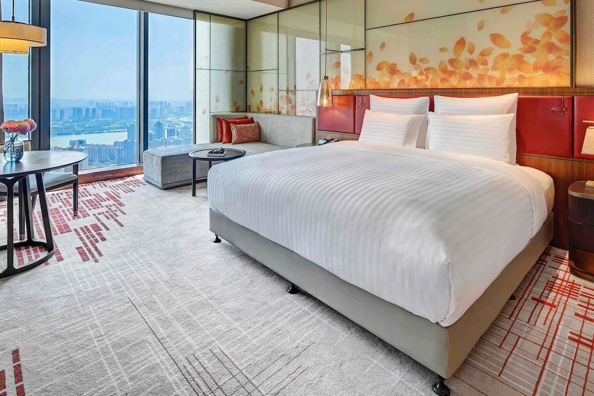 Een moderne hotelkamer met een groot bed, een zithoek met een tafeltje en een uitzicht op de stad door grote ramen.