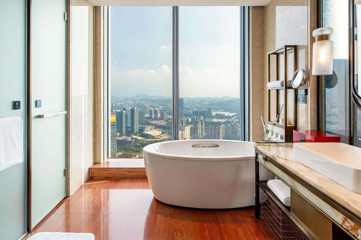 A modern bathroom with a large bathtub, a vanity with a mirror, and a view of a cityscape through large windows.