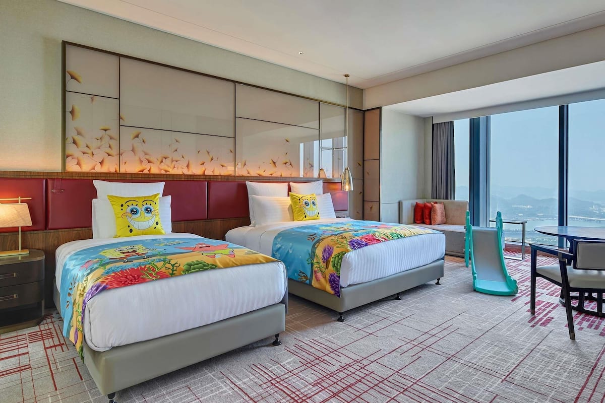 Hotelkamer met twee bedden, een sofa, een eettafel en een groot raam met uitzicht op de stad.