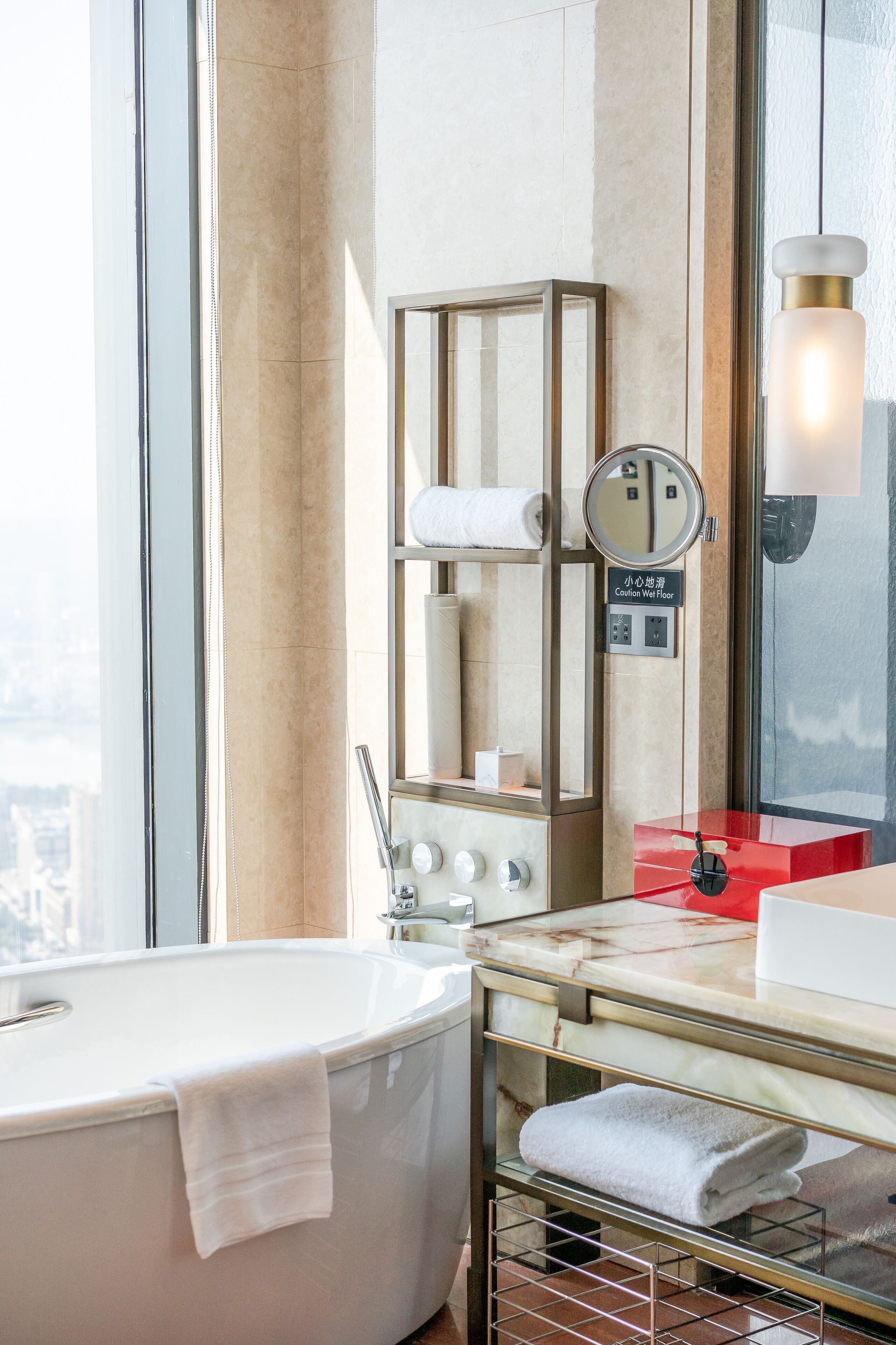 A modern bathroom with a freestanding bathtub, a glass shelf with towels, a vanity with a red countertop, and a large mirror.