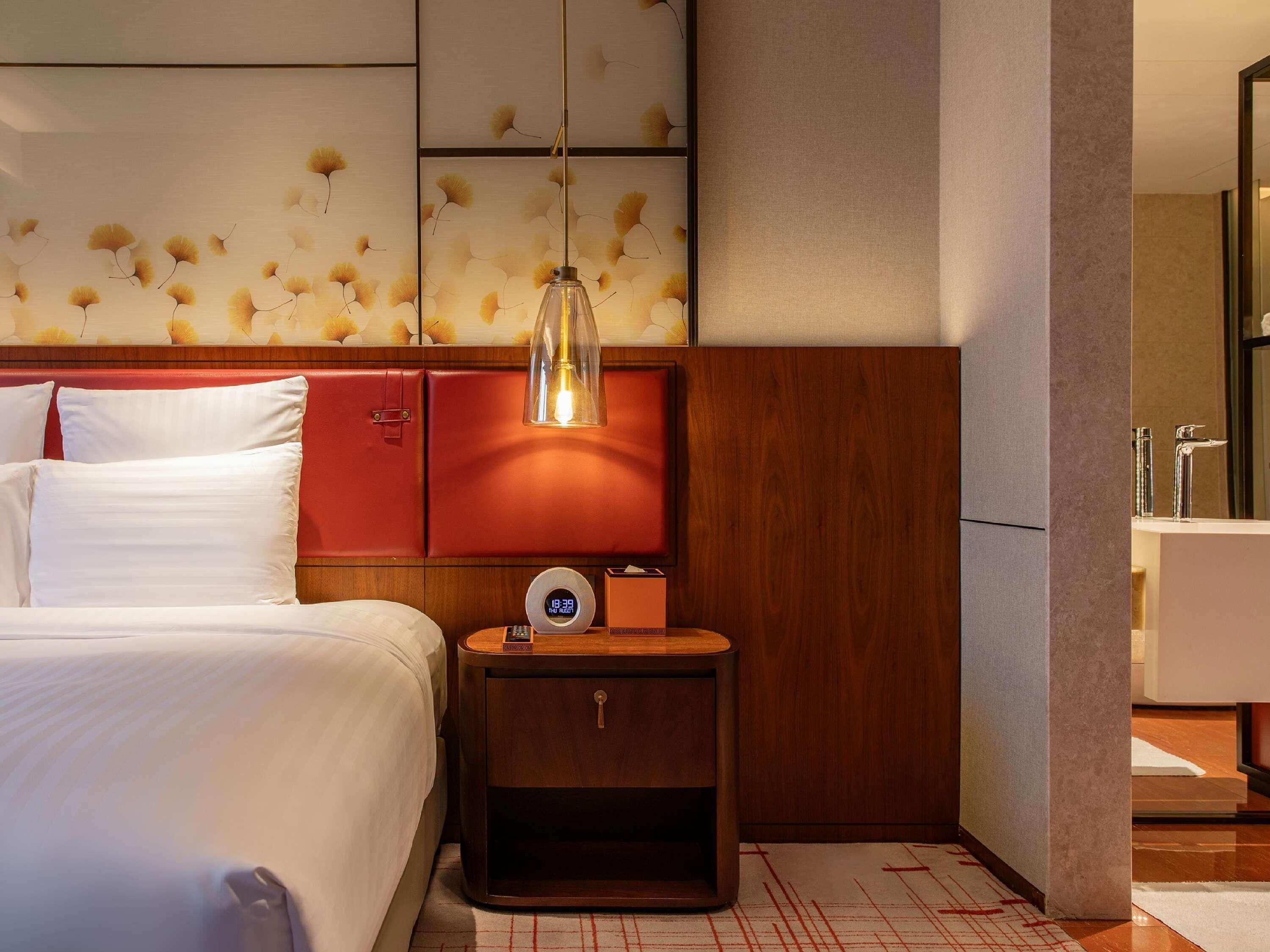 Une chambre d’hôtel avec un lit, une table de chevet avec une horloge, une armoire rouge et un luminaire suspendu.