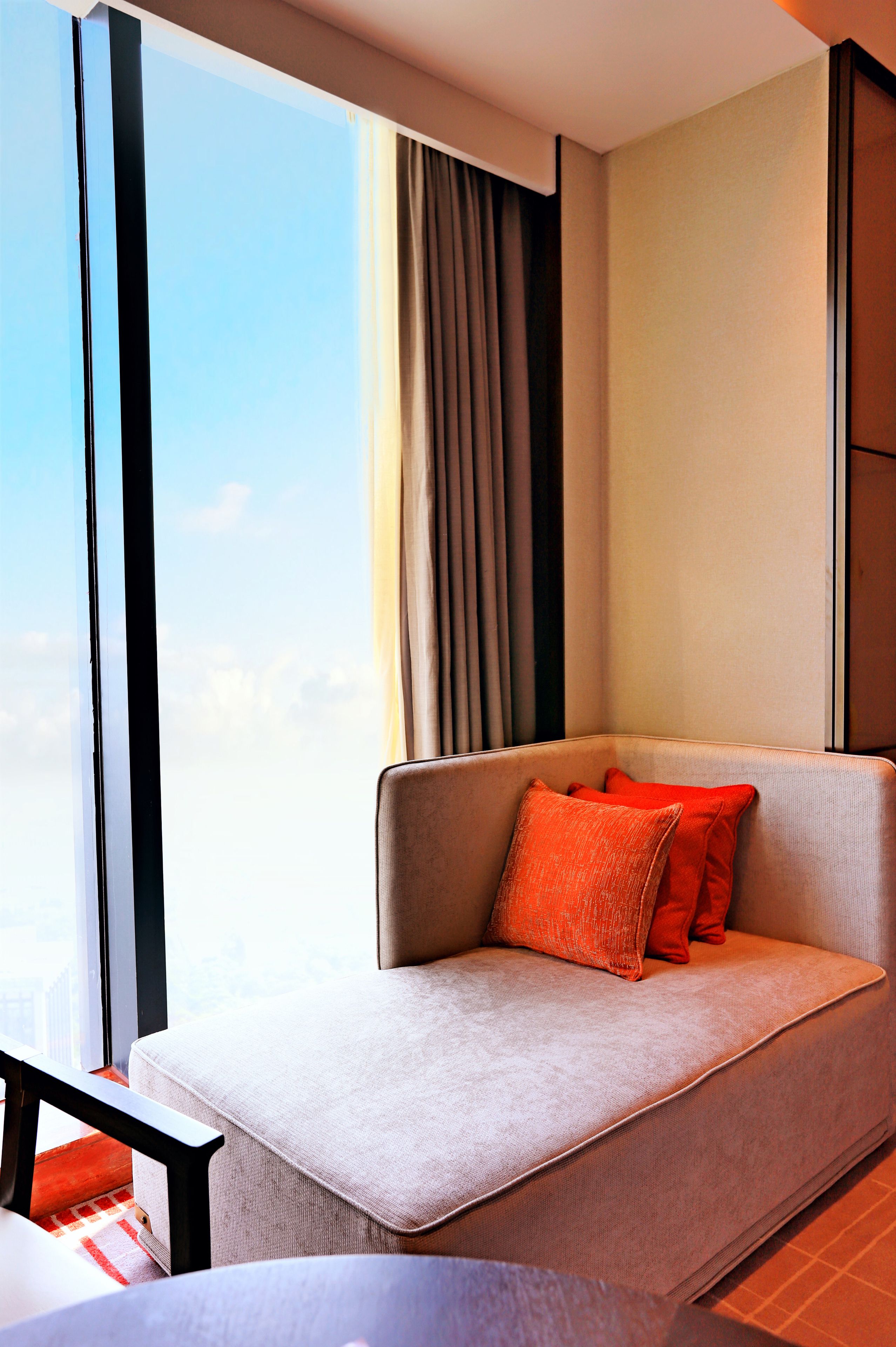 Een moderne hotelkamer met een groot raam, een bank met oranje kussens en uitzicht op de lucht.