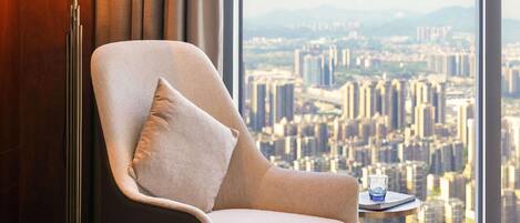 Un sillón moderno con un cojín, una mesita redonda con un vaso, y vistas de un paisaje urbano a través de amplios ventanales.