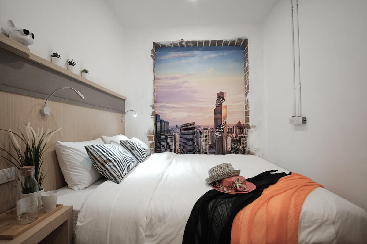 A bedroom with a cityscape tapestry, a wooden headboard, a bedside table with a lamp, and a nightstand with a plant.