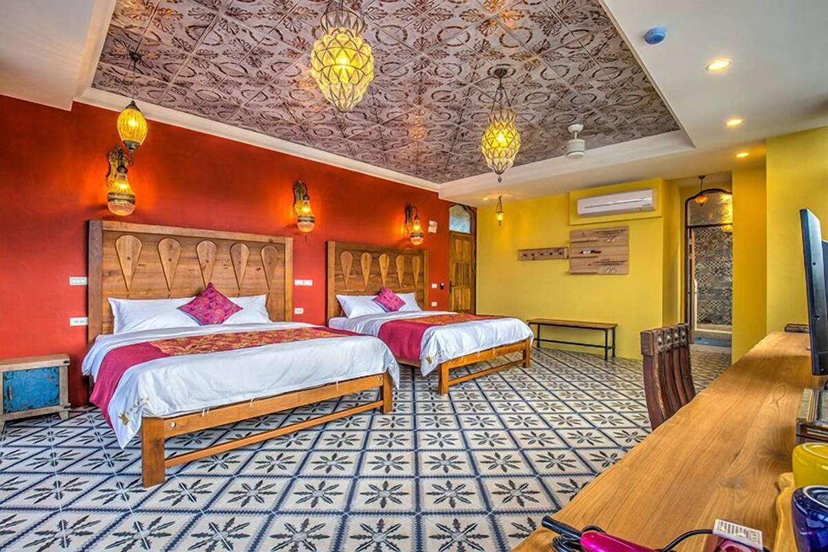 Une chambre d’hôtel avec deux lits, une tête de lit en bois, des carreaux de sol à motifs, une table à manger en bois et un plafond orné de motifs complexes.