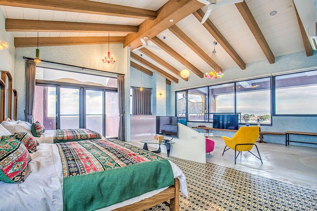 Une pièce spacieuse avec un grand lit, un bureau et une chaise. De grandes fenêtres offrent une vue sur l’extérieur.