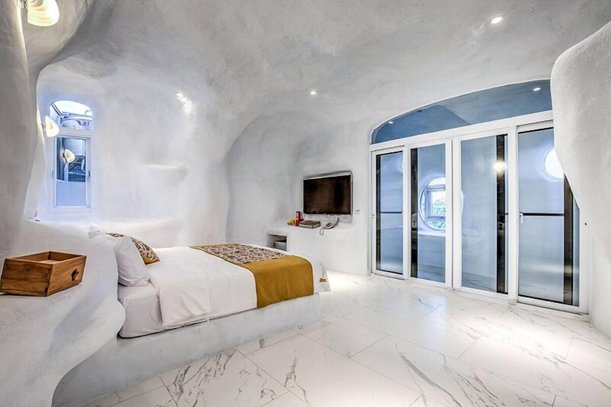 Une chambre moderne avec un plafond incurvé, un lit doté d’une commode en bois, une télévision et une porte-fenêtre coulissante donnant sur un balcon.