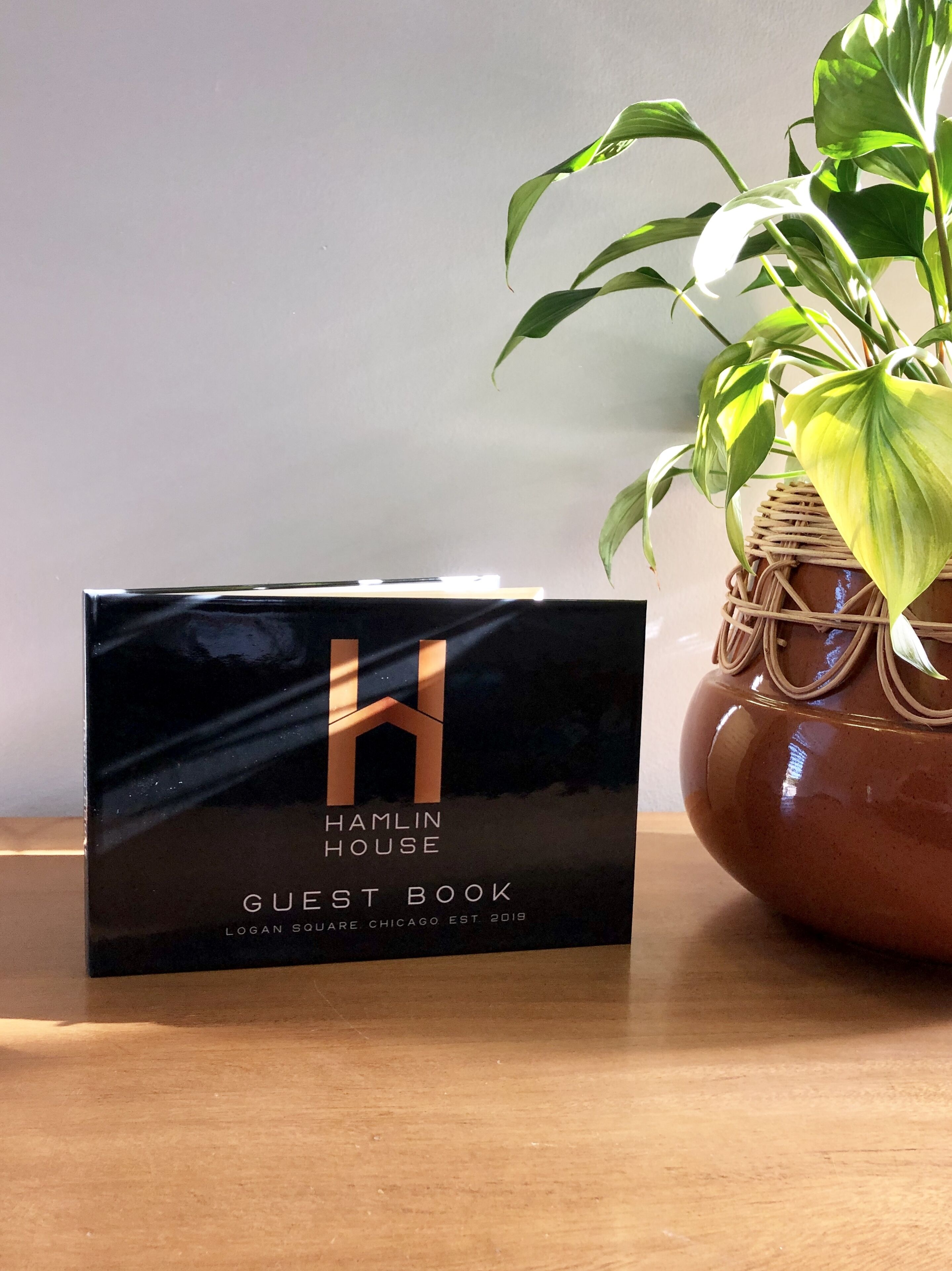 A black guest book with the text "HAMLIN HOUSE LOGAN SQUARE CHICAGO EST 2019" on a wooden table next to a potted plant.