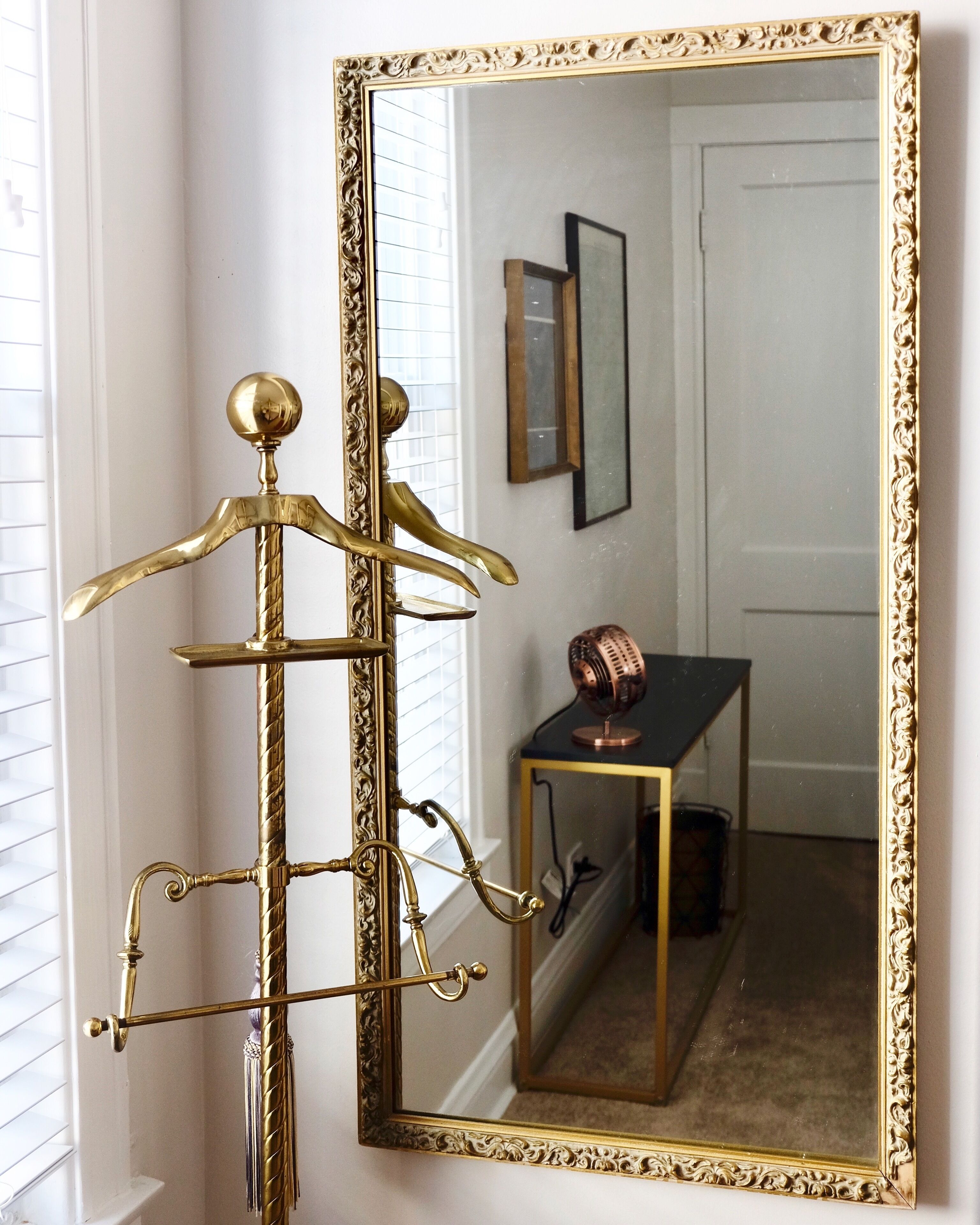 A room with a large ornate mirror, a brass coat rack, and a small table with a decorative fan.