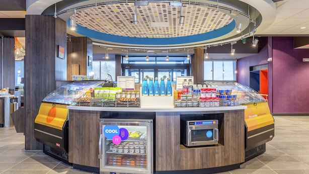 Un quiosco moderno de autoservicio con un expositor refrigerado, una máquina de café y una variedad de productos de snack.
