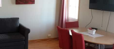 Una habitación con un sofá de color gris oscuro, una mesita redonda con dos sillas rojas, un mantel blanco y un cuadro abstracto rojo en la pared.