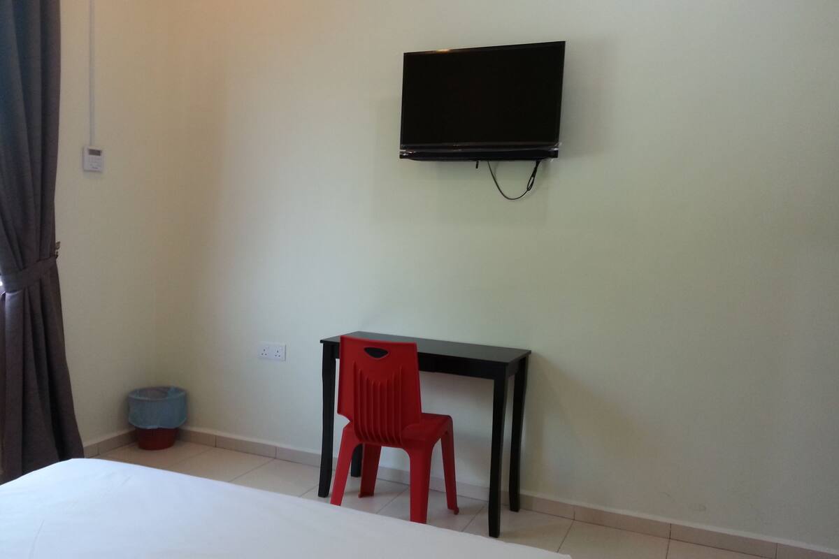 A room with a bed, a desk, a red chair, a television on the wall, and a small trash bin.