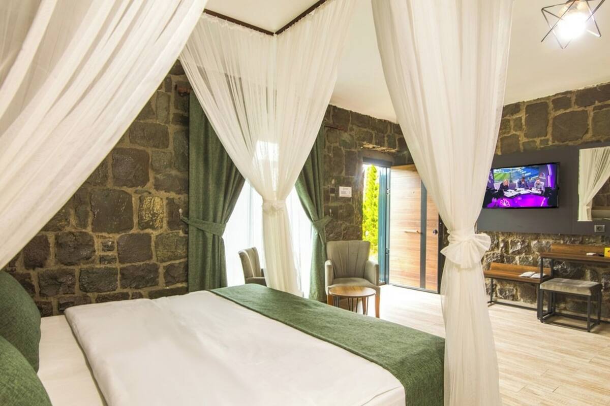 A bedroom with a stone wall, a bed with white linens, a green chair, a TV, and a wooden bench.