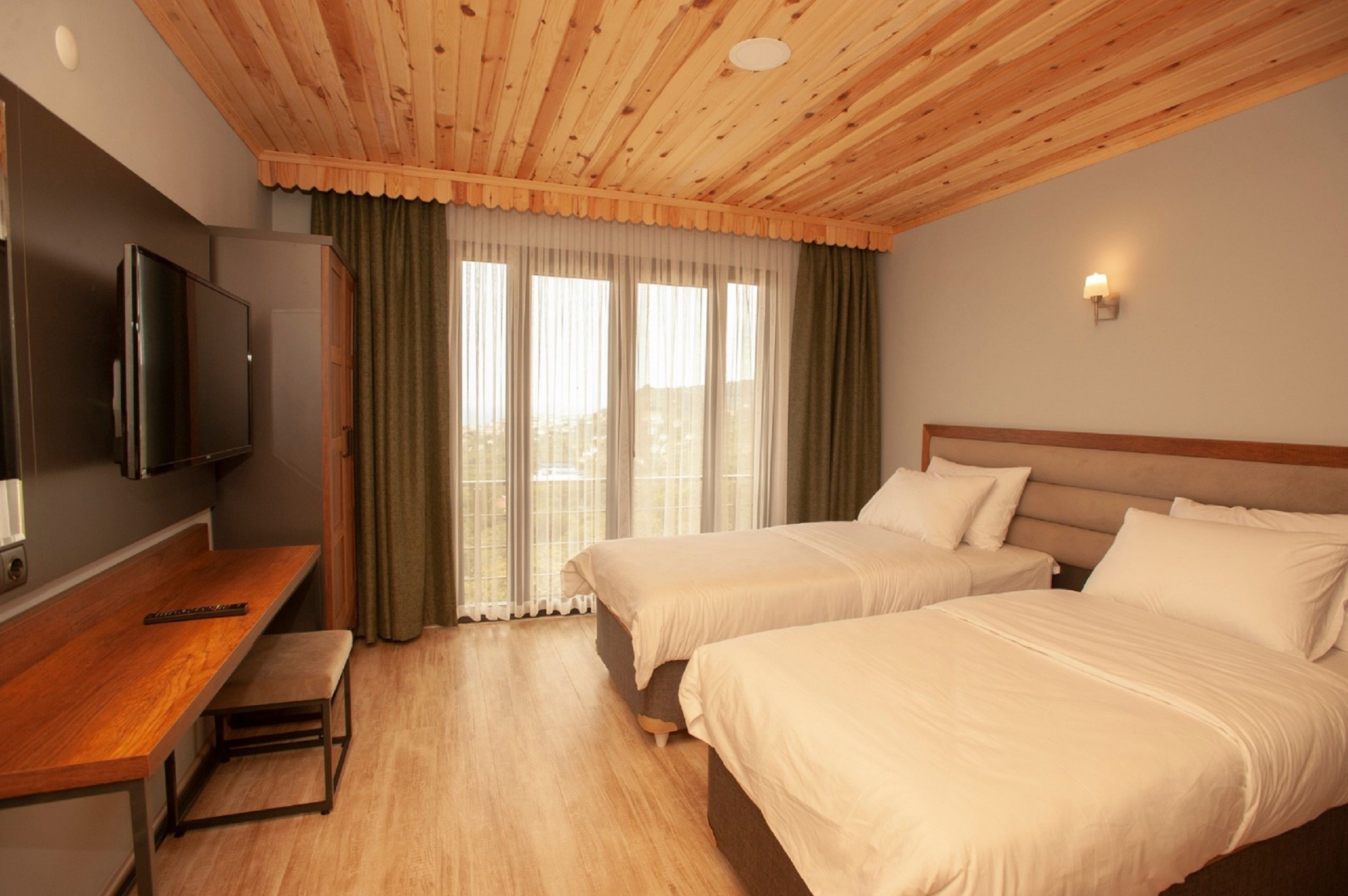 A hotel room with two beds, a wooden ceiling, a TV, a desk, and a window with curtains.