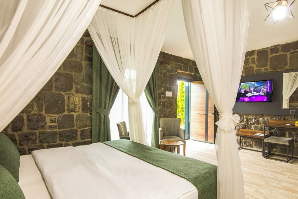 Un dormitorio con pared de piedra, cama con ropa blanca, una silla verde, televisor y un banco de madera.