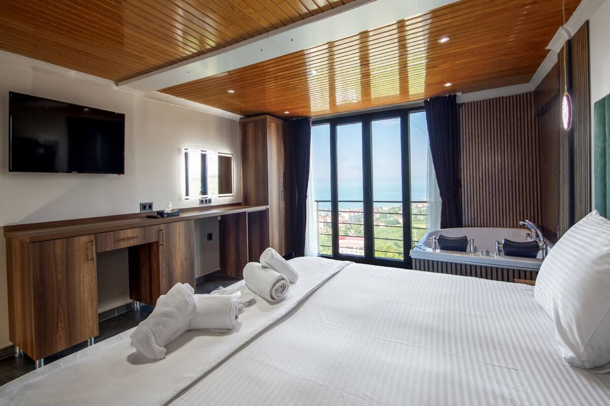 A modern hotel room with a wooden ceiling, a large bed, a TV, a desk, and a view of the sea.