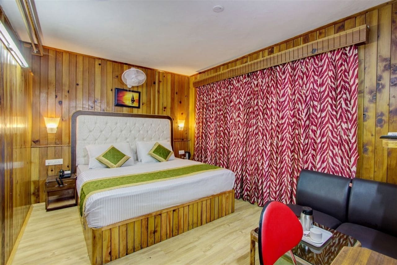 A wooden-paneled room with a bed, a small table with a red chair, and a patterned curtain.