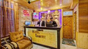 A reception desk with two staff members, a sign indicating CCTV surveillance, and a reception area with a sofa and a wooden wall. - Hotel Samiru (Manali)