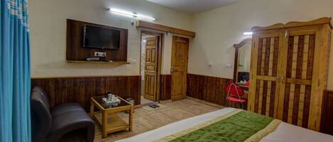 A hotel room with a bed, a TV mounted on the wall, a wooden wardrobe, and a small table with a chair.