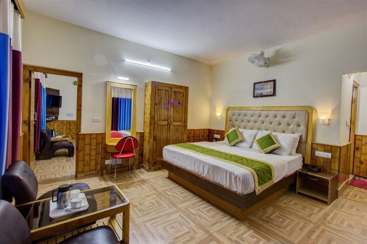 A hotel room with a bed, a small table with a cup and a thermos, a chair, a mirror, a wardrobe, and a ceiling fan.
